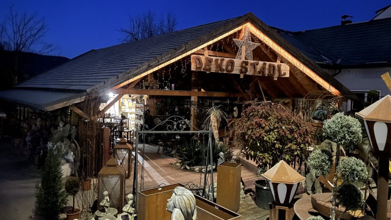 A decoratively illuminated wooden building with the inscription 'Deko Stadl' at night, surrounded by plants and decorative items.