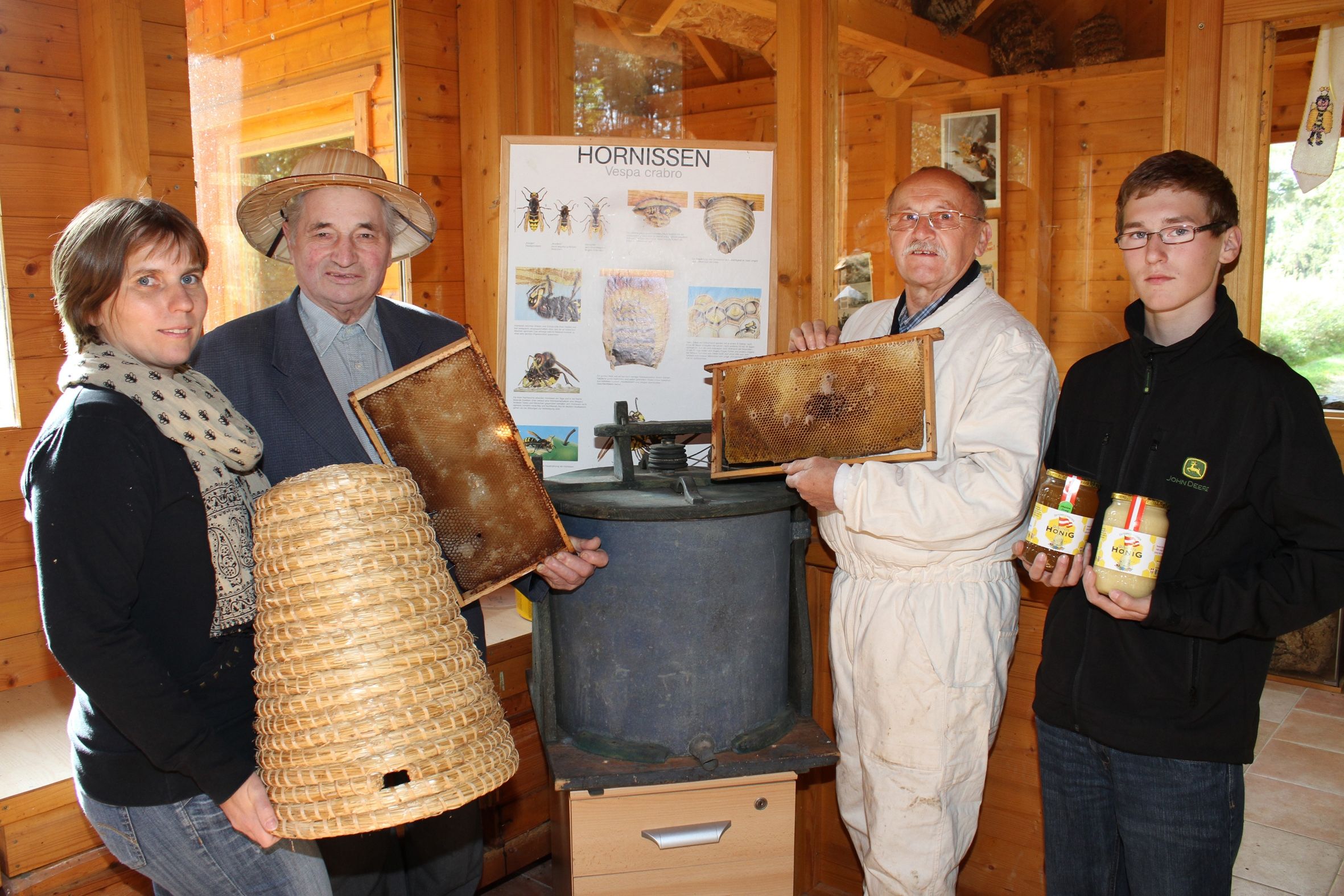 Four people in a wooden room with beekeeping equipment and honey jars.