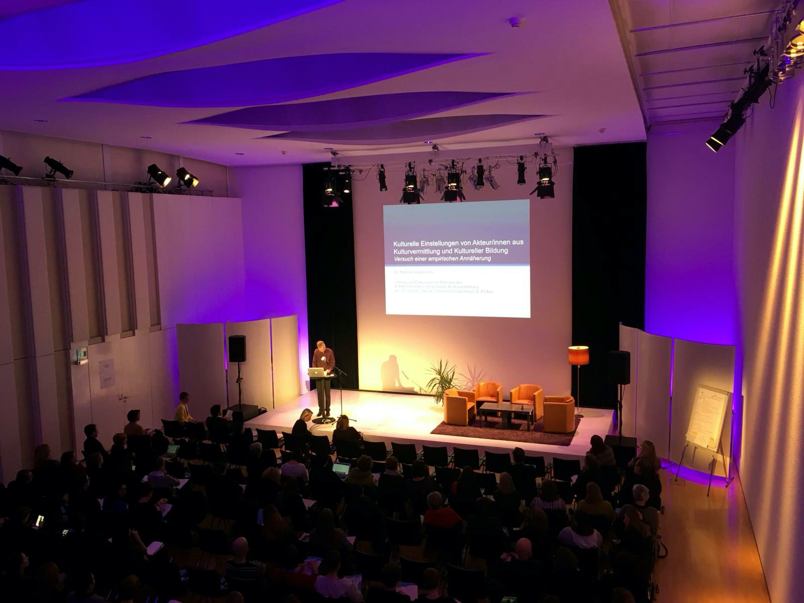 A lecture in a conference room with purple lighting and an audience.