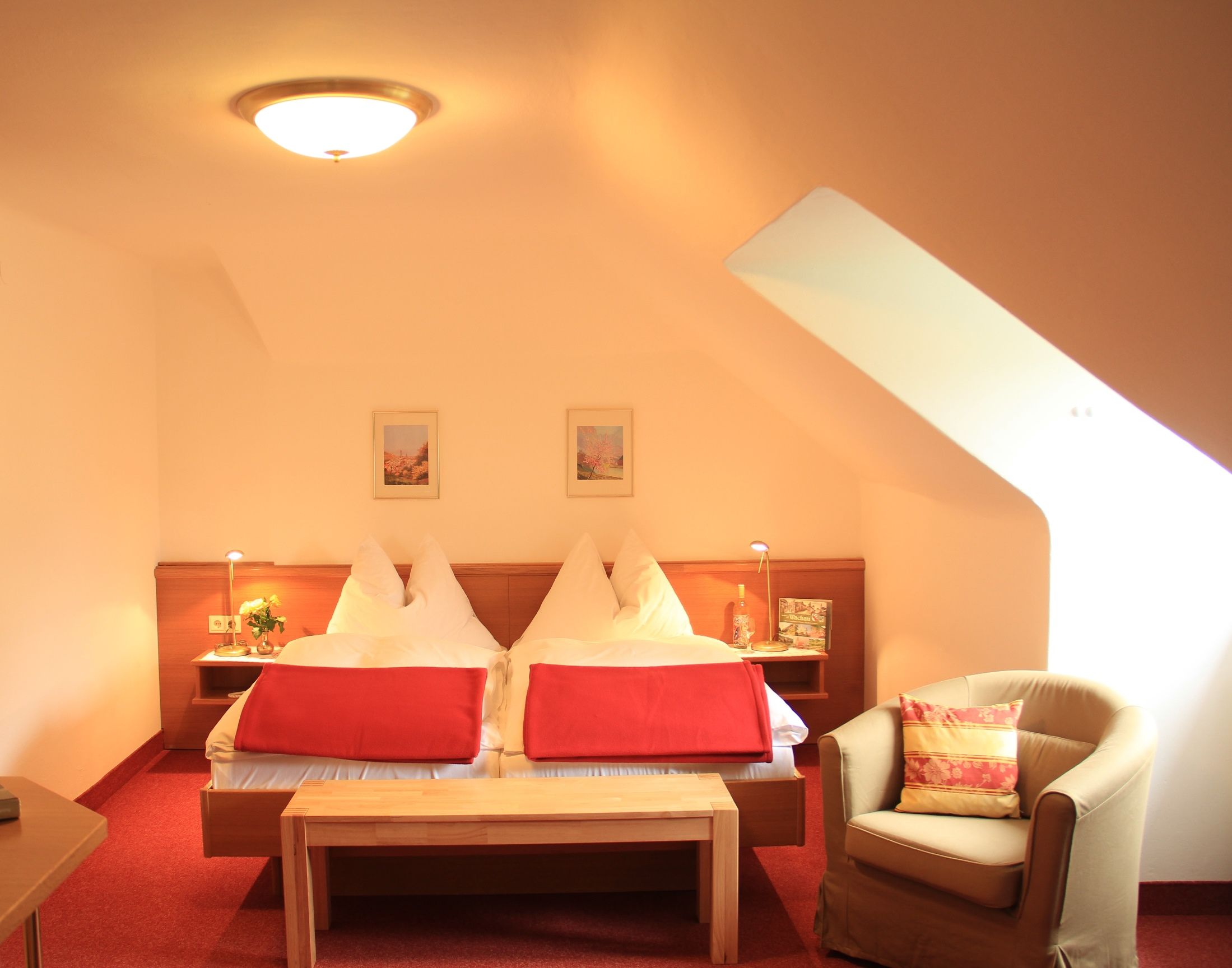 A cozy double room with two beds, red blankets, an armchair and a table under a sloping roof.