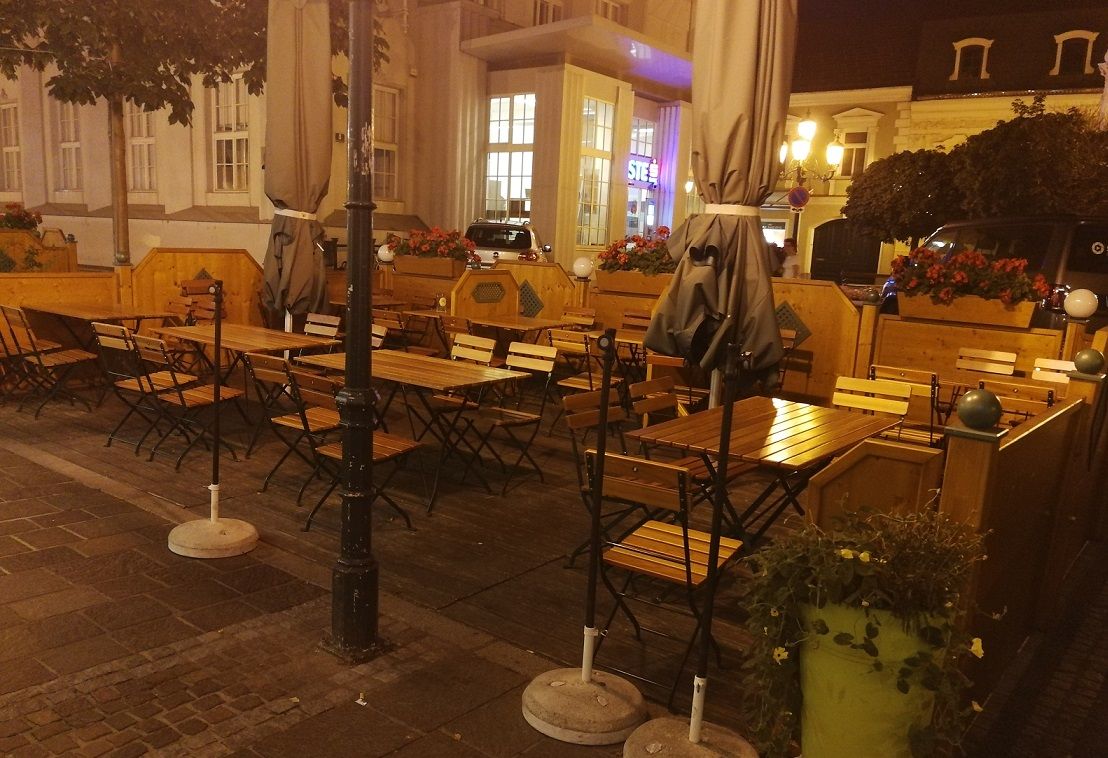 Outdoor area of a restaurant with empty wooden tables and chairs, surrounded by plants and lanterns at night.