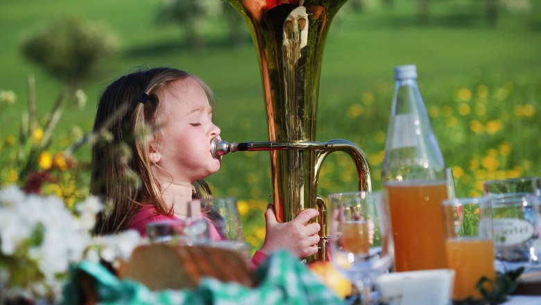 A little girl plays a tuba on a meadow, surrounded by drinks and flowers.