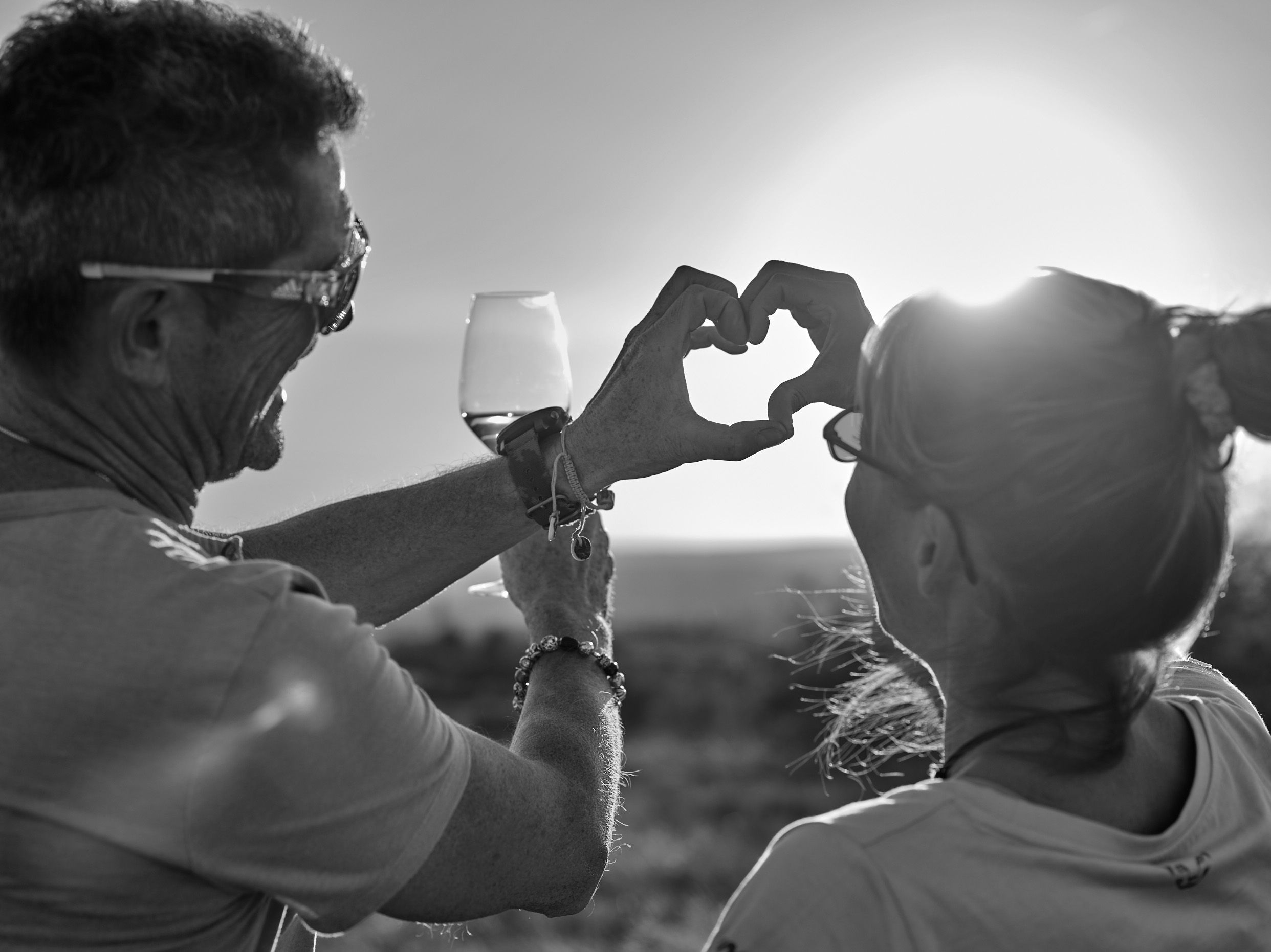 Two people form a heart with their hands in the sunset, one is holding a wine glass.