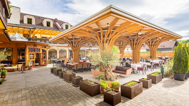 Terrace with wooden roof and tables, surrounded by plants.