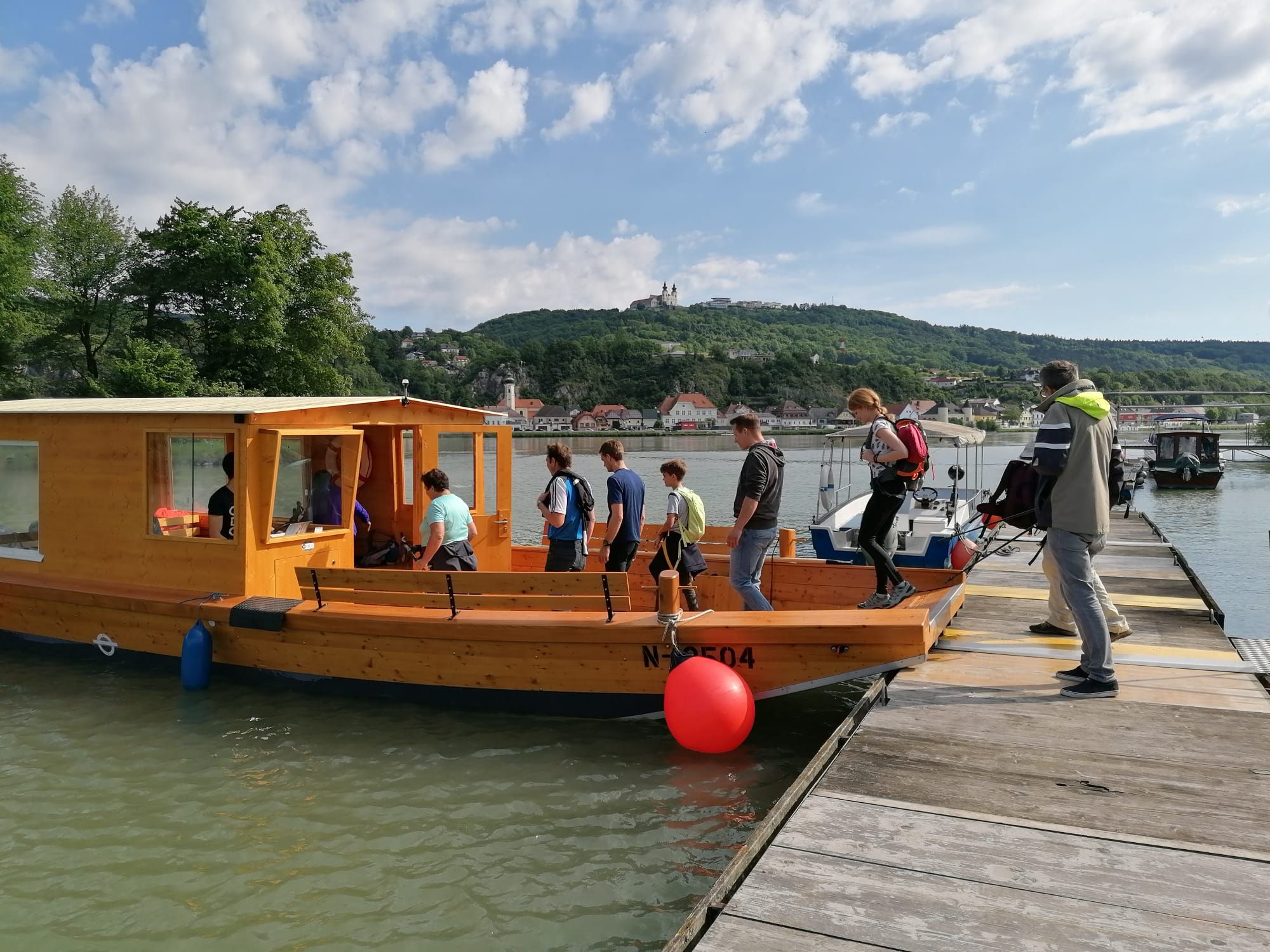 People board a wooden boat on a jetty, in the background a hilly landscape with buildings.
