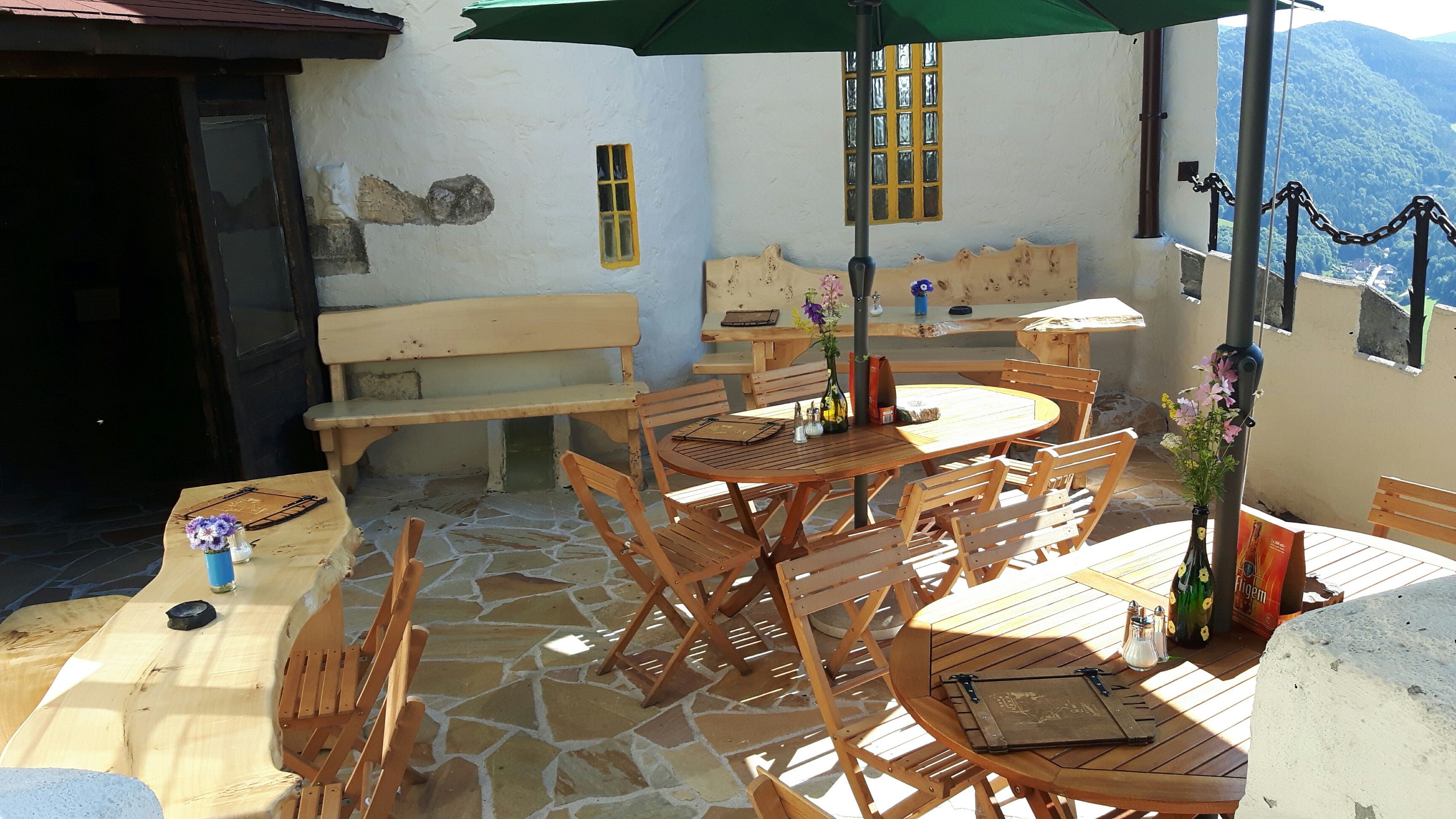 Wooden terrace with tables, chairs and parasol, surrounded by a stone wall with mountain views.