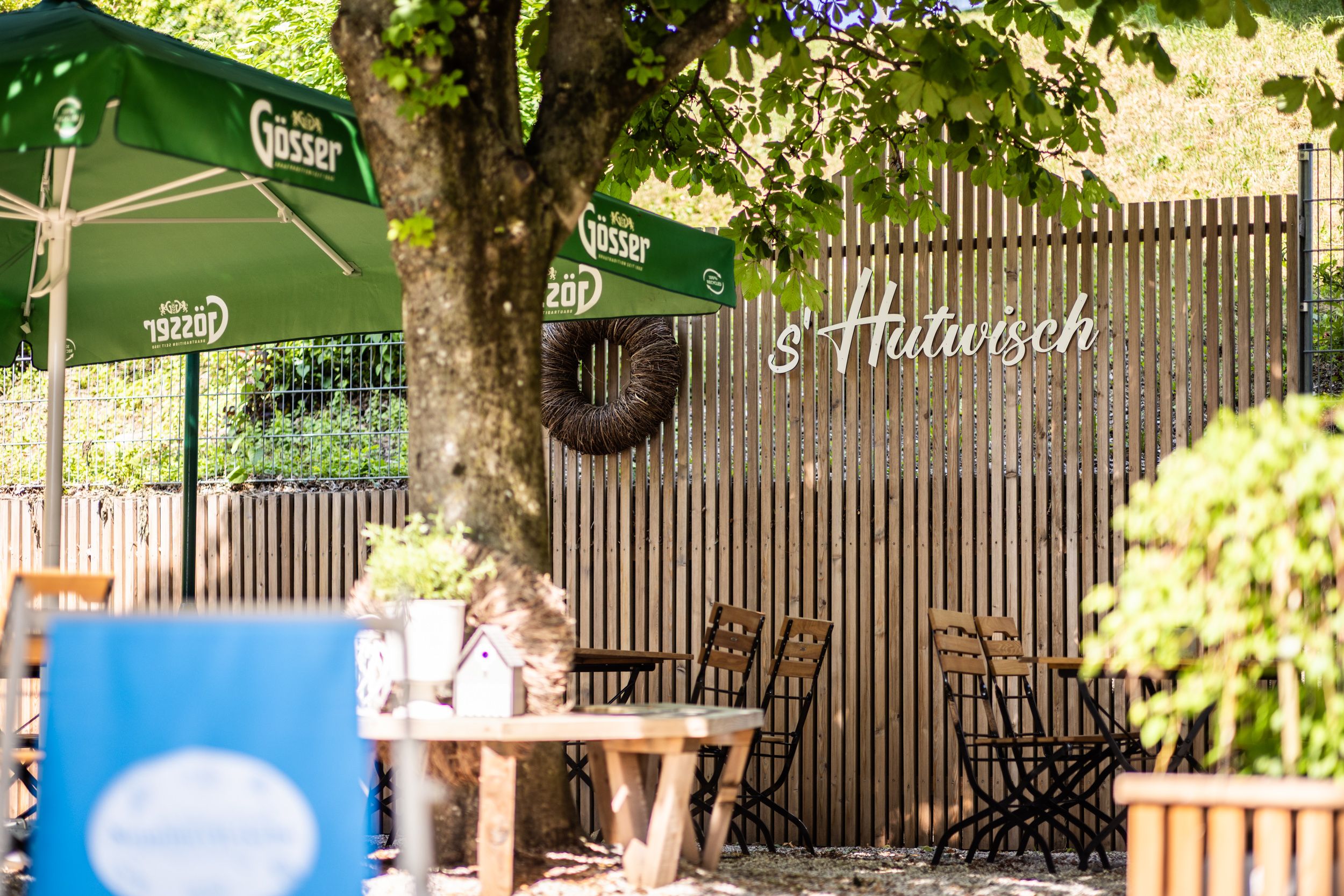 A guest garden with wooden tables and chairs, green parasols and a wooden fence with the inscription 's'Hutwisch'. In the middle is a tree surrounded by a bench.