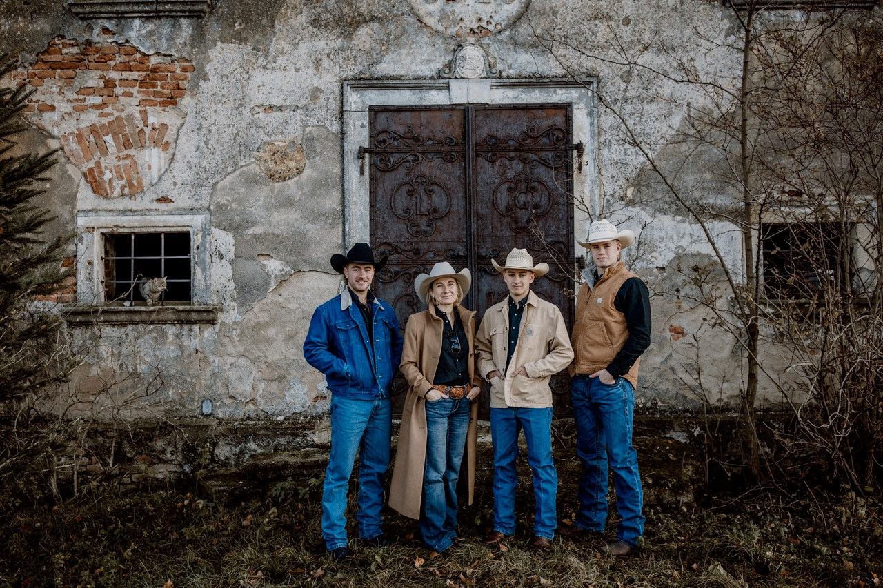 Four people in cowboy clothing stand in front of an old, dilapidated wall with a large metal door.