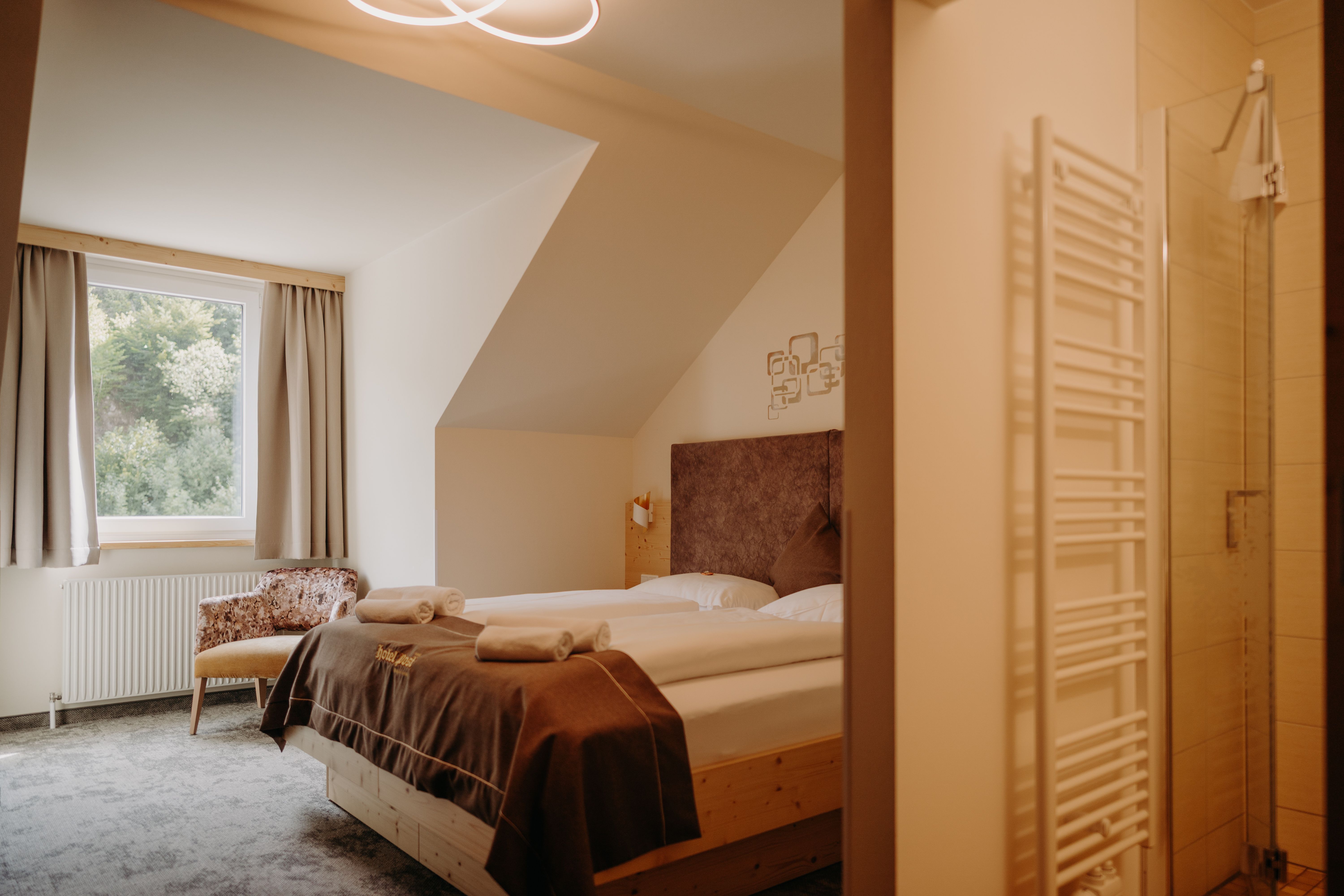 A cozy hotel room with a double bed made of light-colored wood, a window with curtains, and an armchair. In the foreground, you can see into the bathroom. The room has a sloped ceiling on the side with the window.