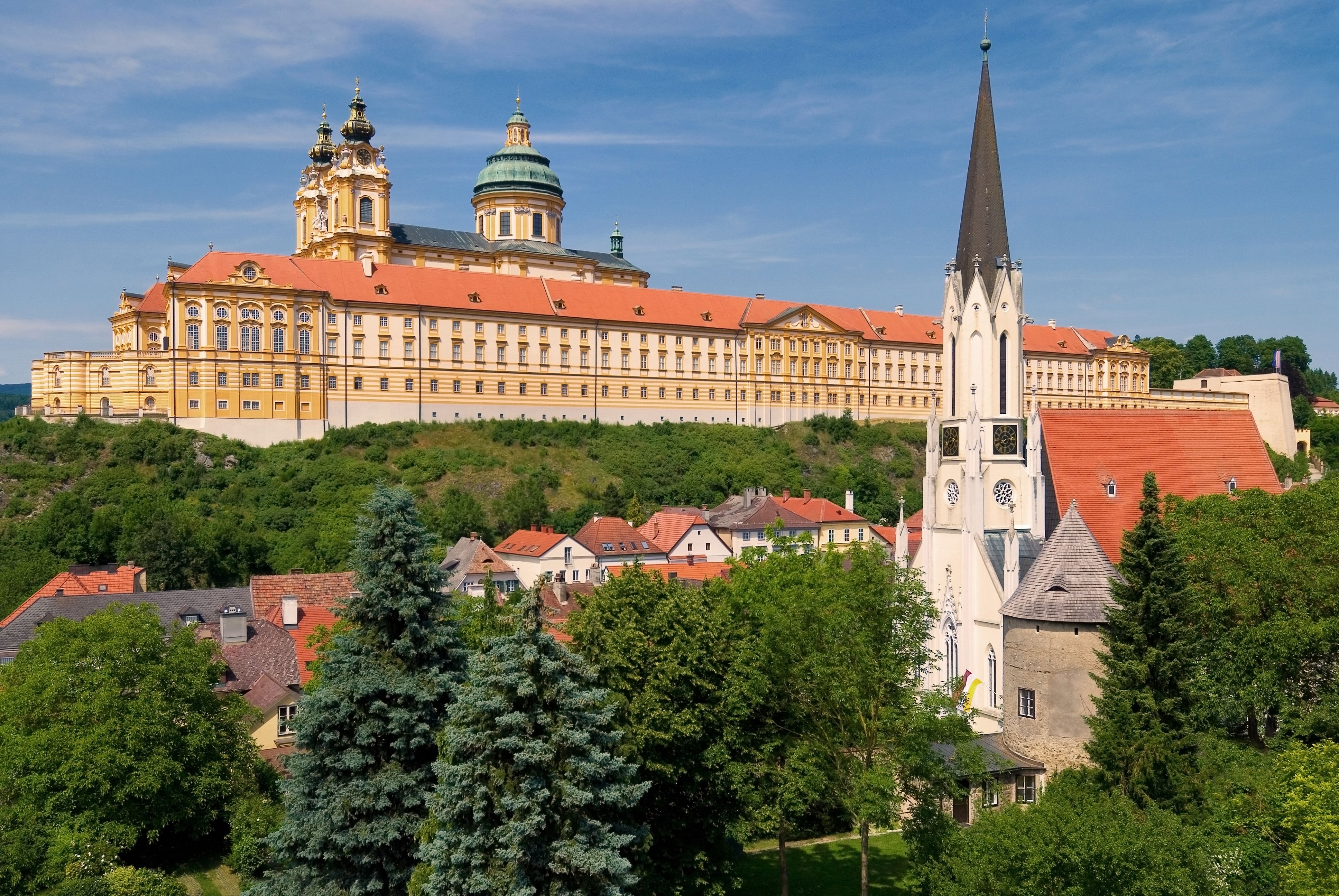 View of Melk Abbey and the parish church in Melk, surrounded by green countryside and blue sky.