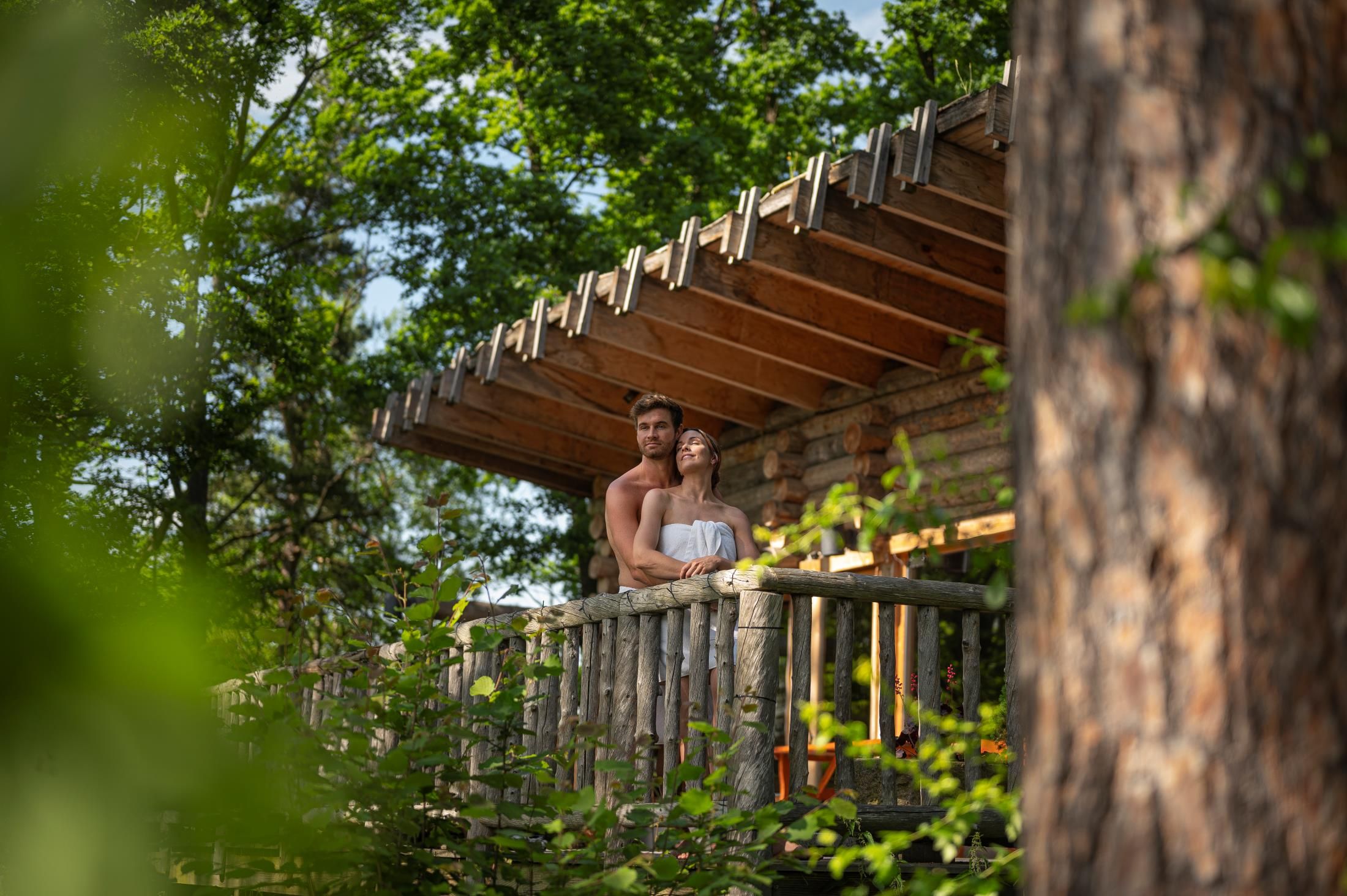 A couple stands on a wooden balcony in a forest, surrounded by trees.