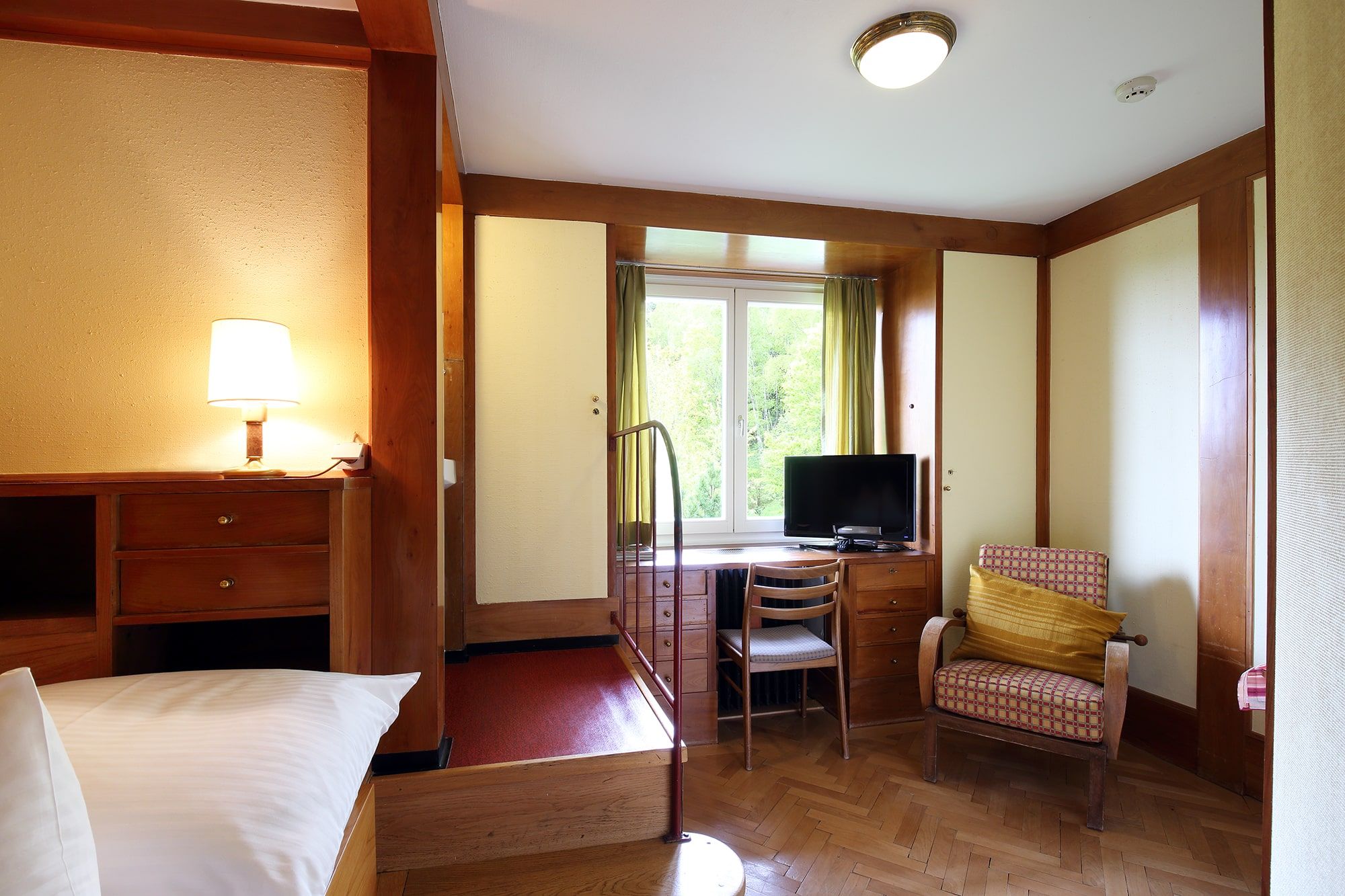 A cozy hotel room with wooden furniture, a TV, an armchair and a window with a view of the greenery.