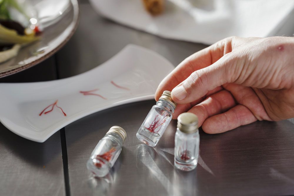 Her hand holds a small glass vial with saffron threads, next to it two other vials and a plate with saffron.
