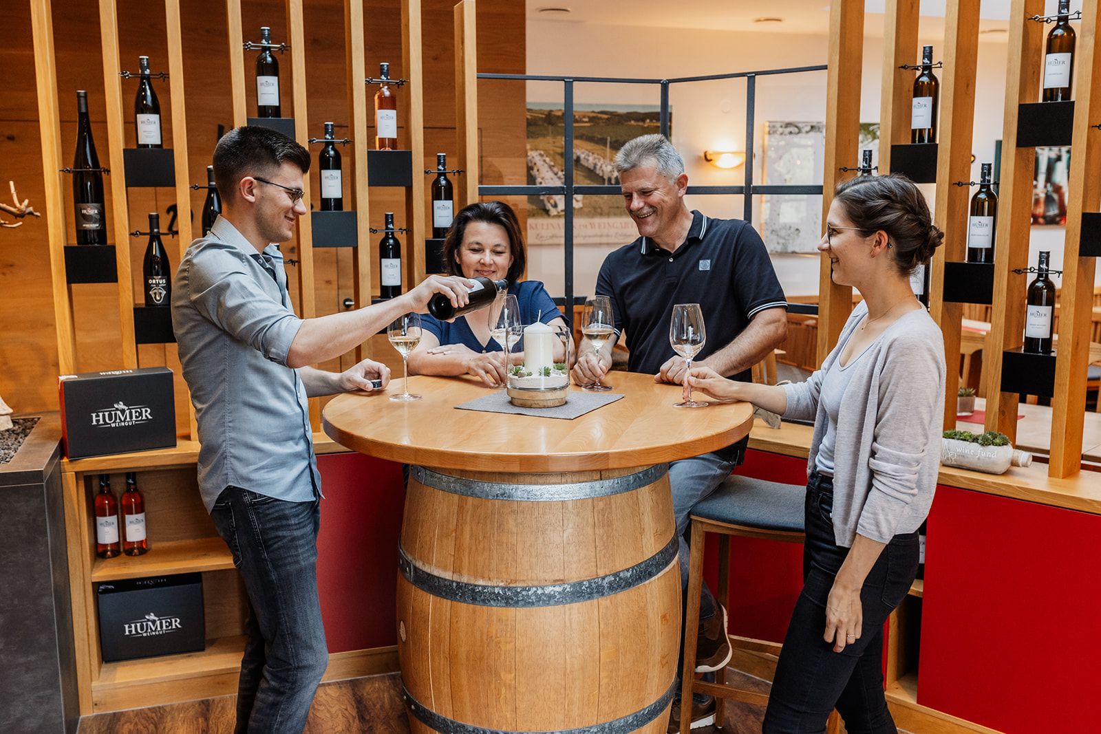 Four people stand around a round table made from a wine barrel, while one person pours wine.