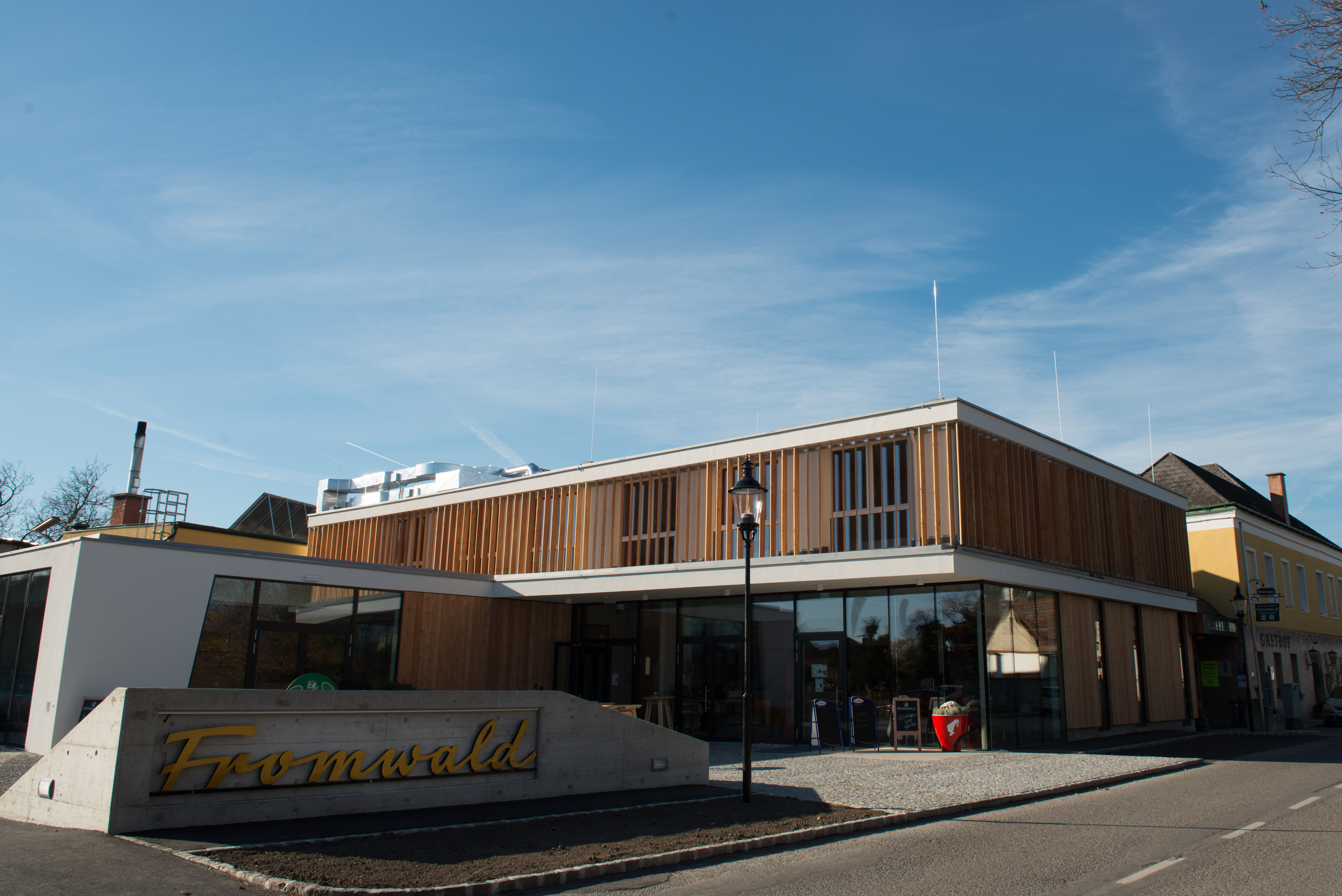 Modern building with wooden façade and glass front, in front of it a sign with the inscription 'Fromwald'.