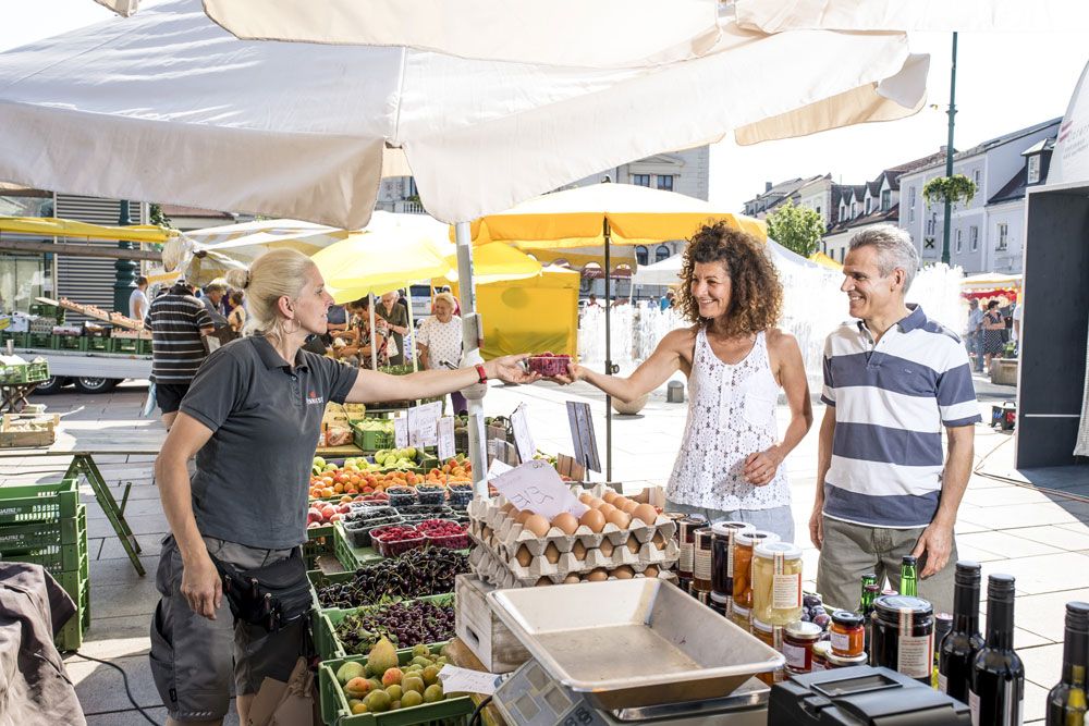 People buy fresh produce at a market with stalls and umbrellas.