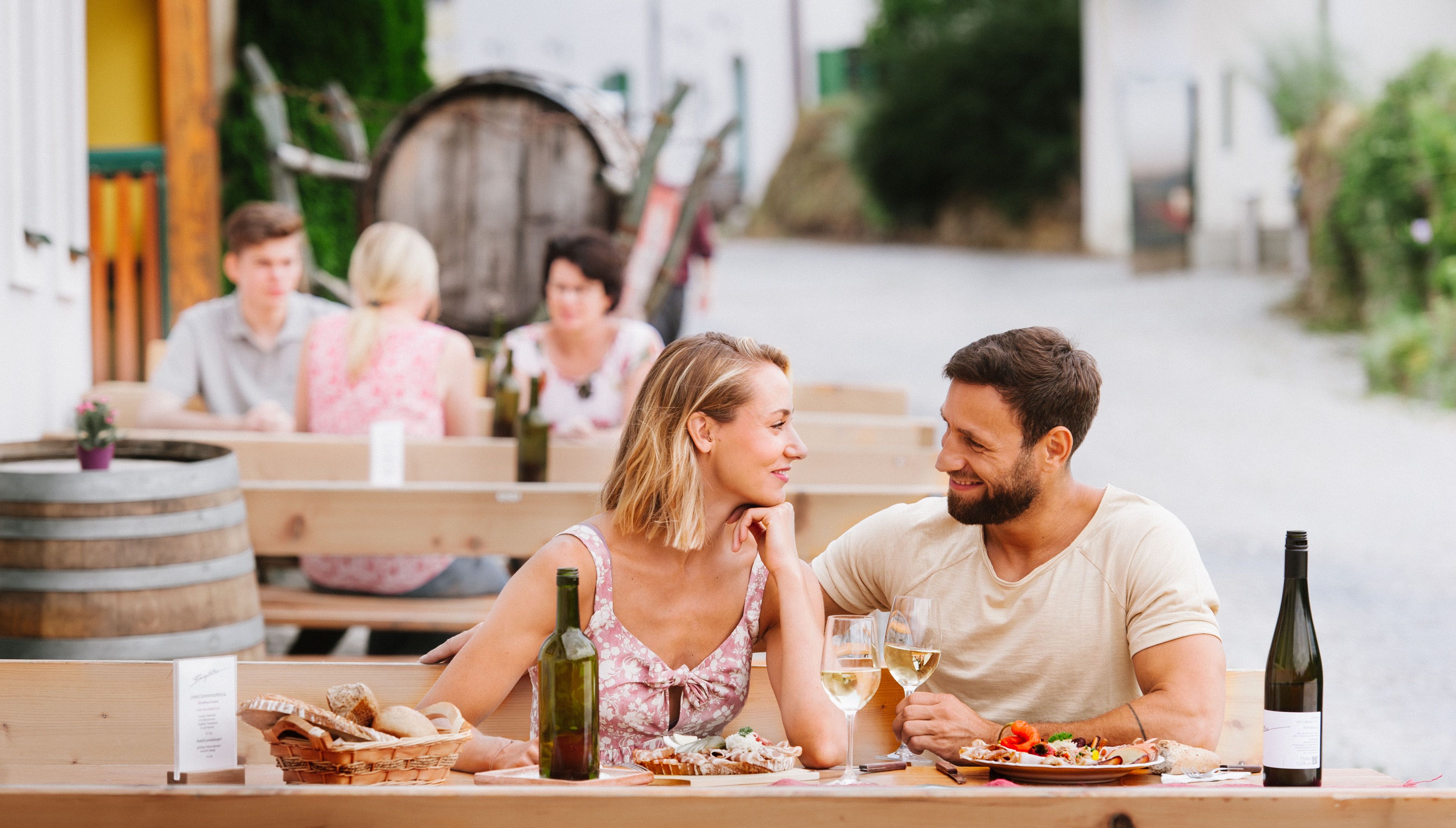A couple is sitting at an outdoor table, enjoying wine and food. Other guests and a wine barrel can be seen in the background.