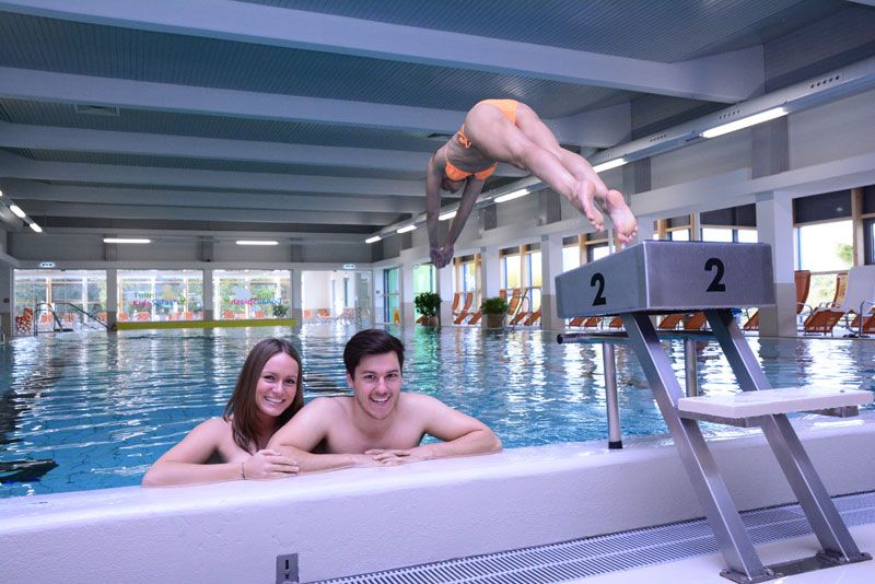 Two people in the swimming pool, one person jumps from the starting block into the water.