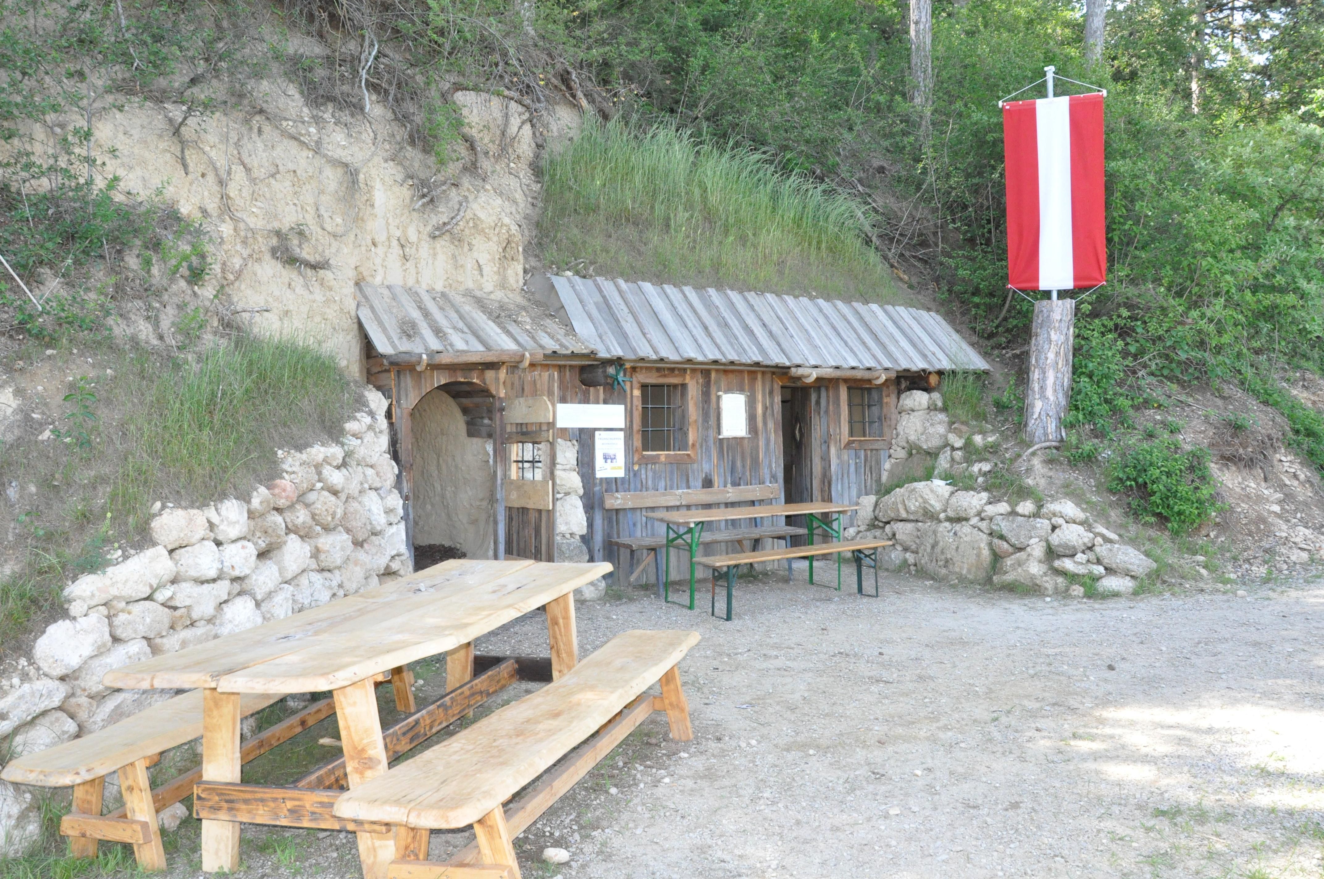 A rustic wooden hut with a table and benches in front of it, built into a hill. A red and white flag hangs from a tree.
