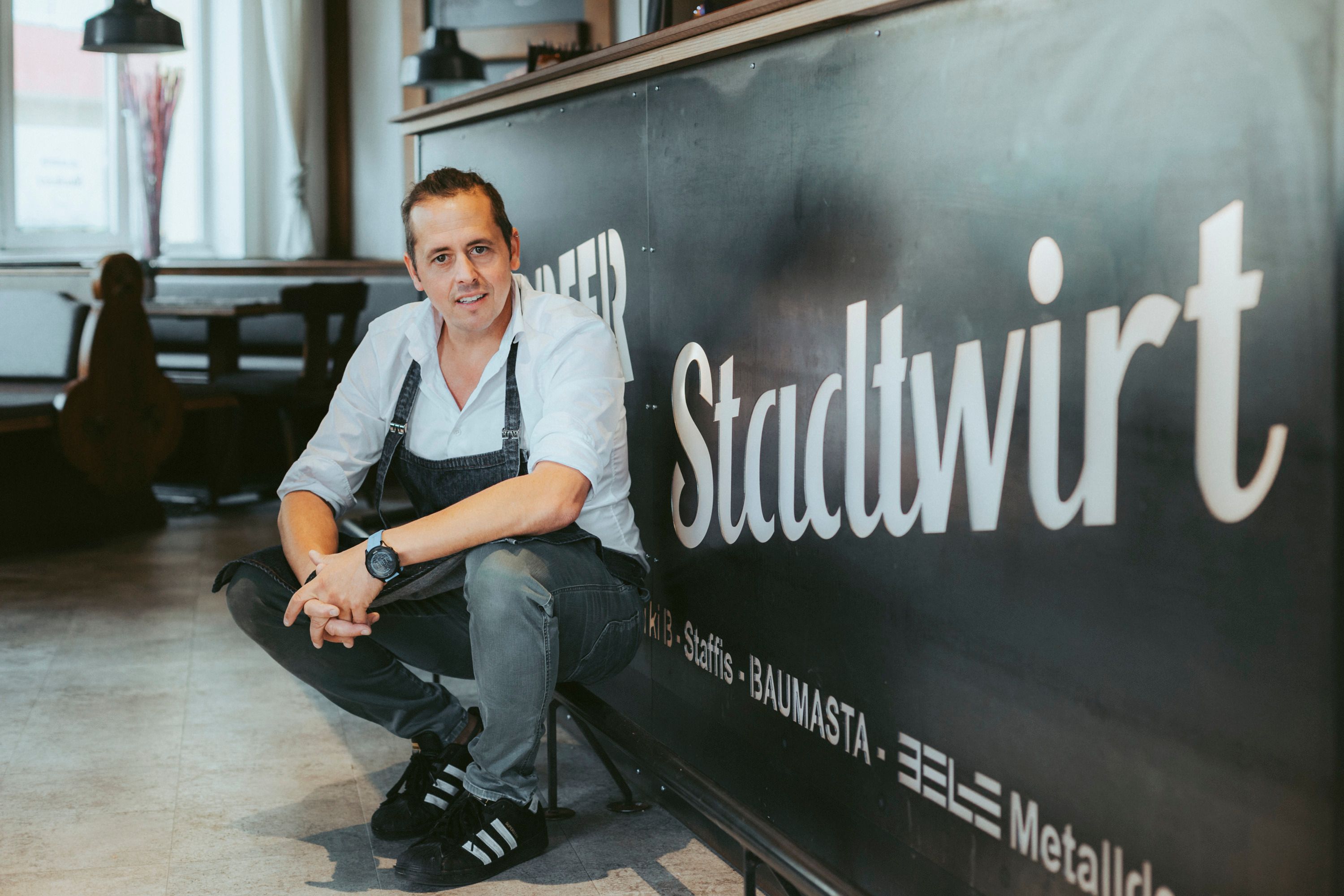 A man in work clothes squats in front of a sign that reads 'Stadtwirt'.