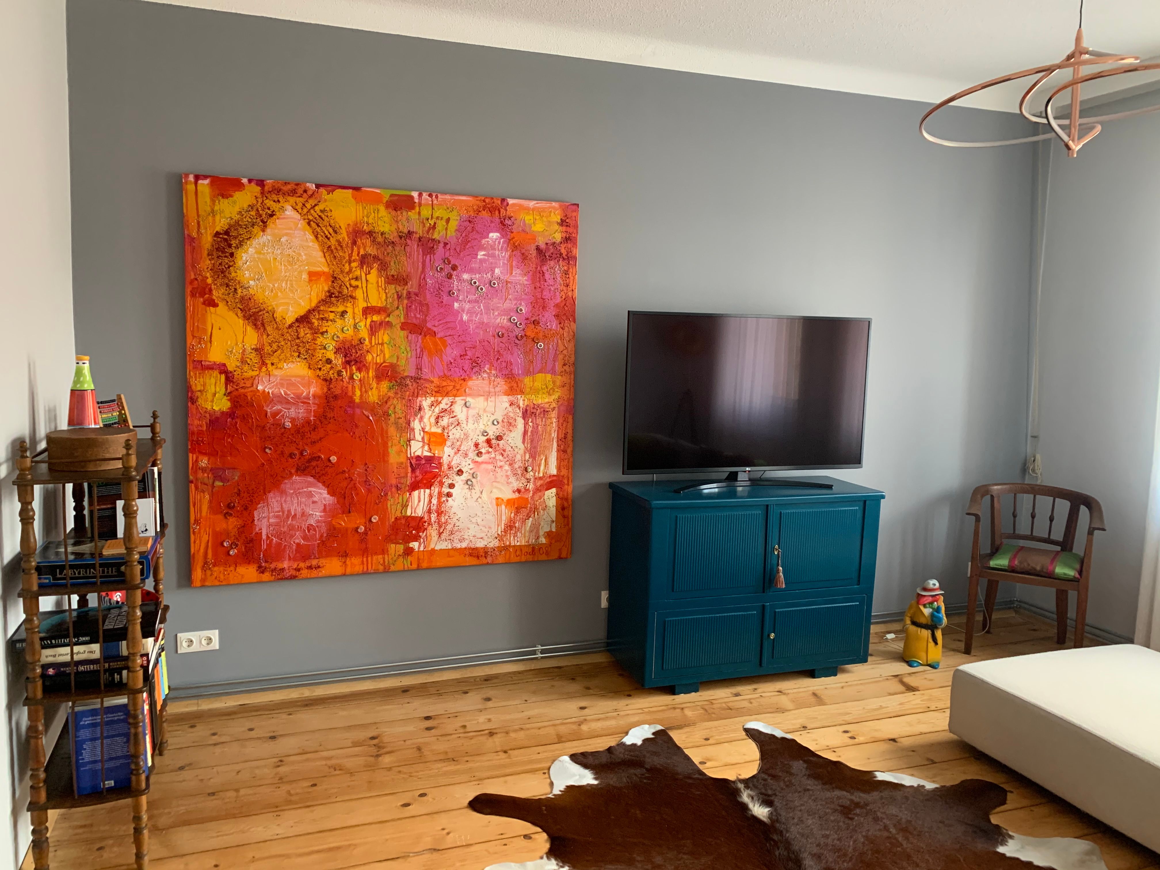 Living room with large colorful painting, TV on blue cabinet, wooden floor and cowhide carpet.