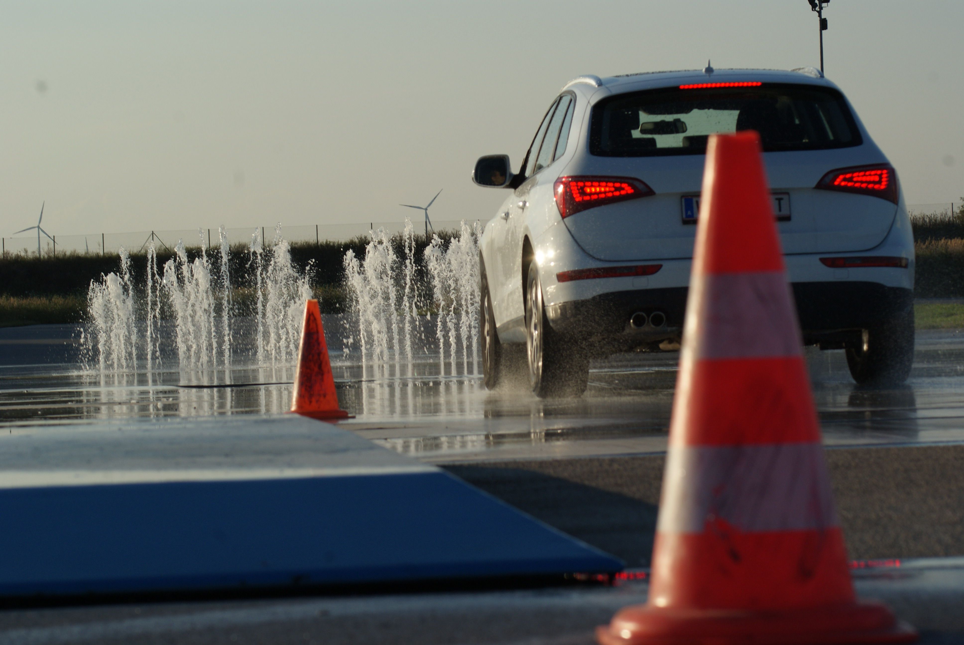 A car drives on a wet road with water fountains and traffic cones.