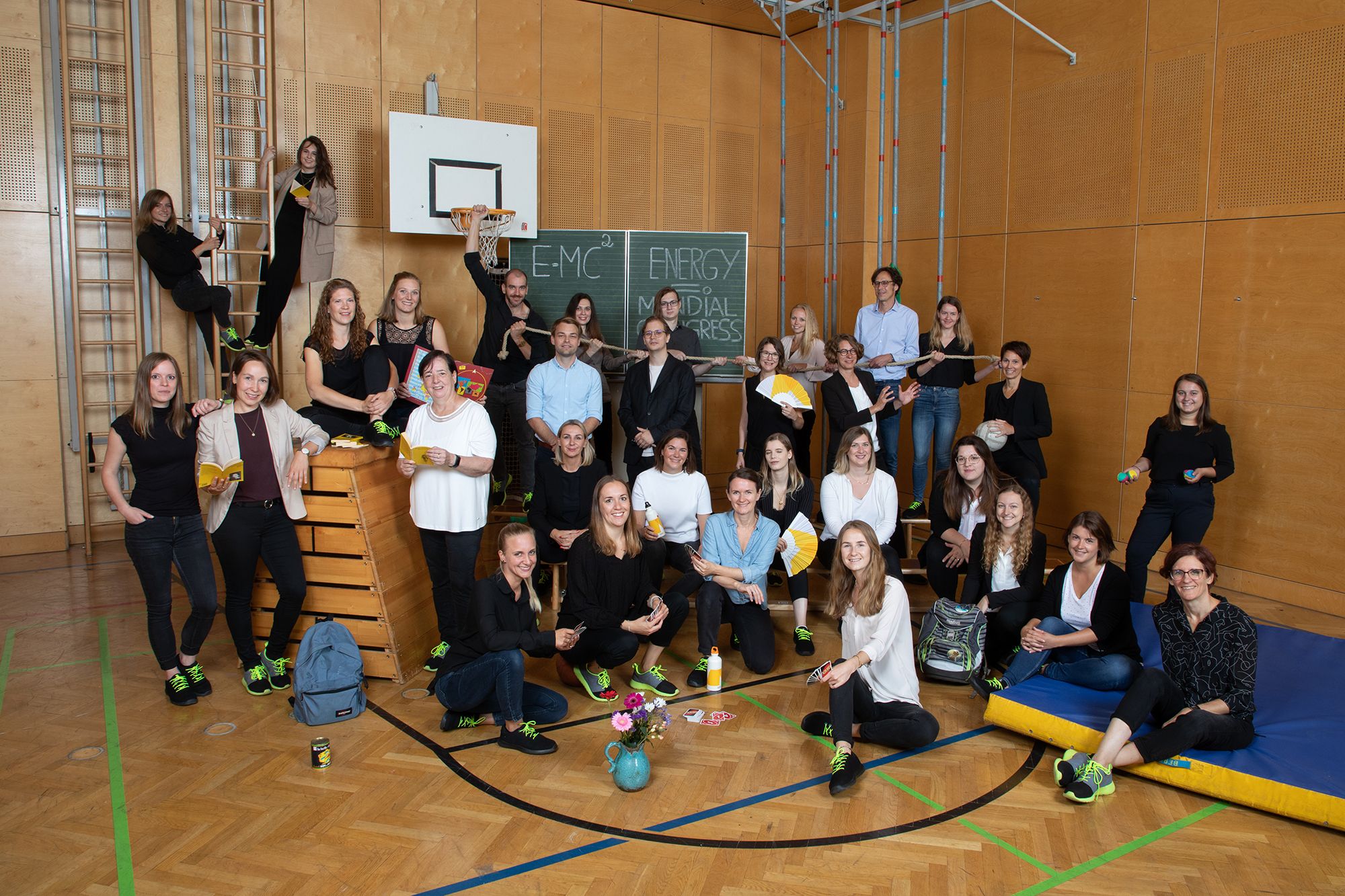 Group of people in a gym with sports equipment and blackboard in the background.