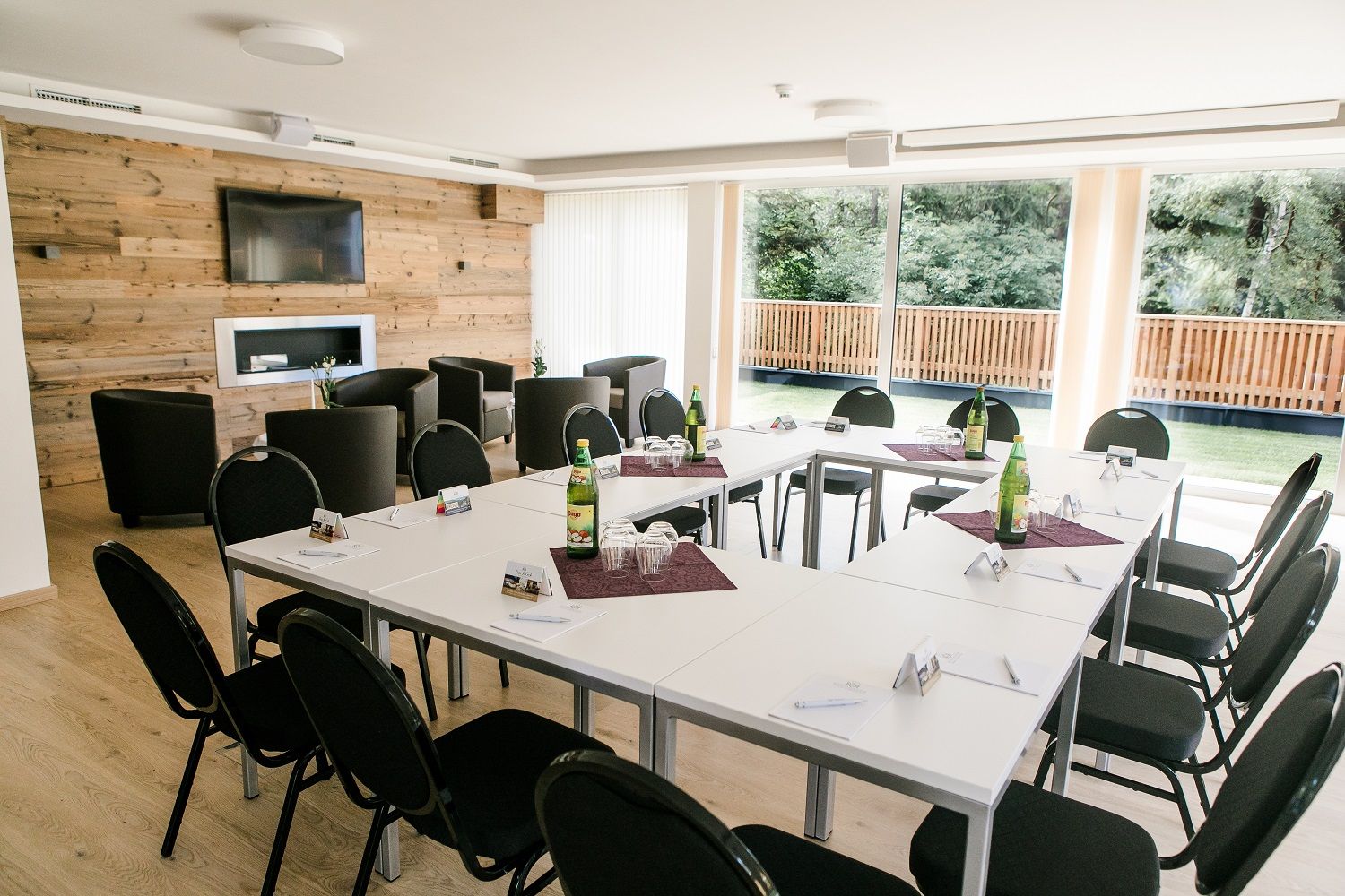 Conference room with U-shaped table arrangement, chairs, wooden wall, TV and large windows with a view of the greenery.