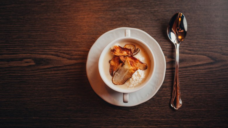 A cup of Jerusalem artichoke soup with chips on a wooden table, a spoon next to it.