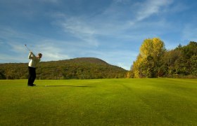 Golfer hits ball on a green golf course with wooded hills in the background.