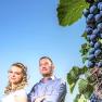 A man and a woman stand in front of a blue sky next to a vine with grapes.