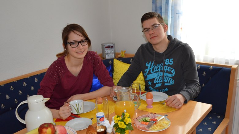 Two people sit at a table with breakfast, including bread rolls, fruit and juice.