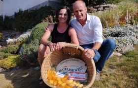 A woman and a man sit smiling in a garden, holding a basket of potatoes and eggs.
