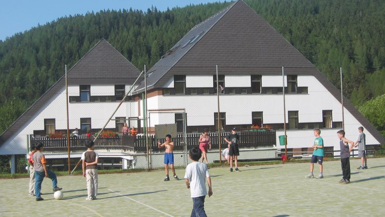 Children play soccer on a sports field in front of a hotel in a wooded area.