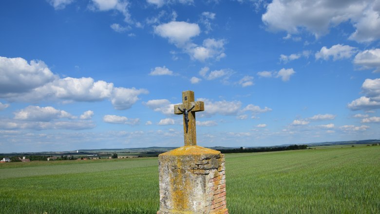 A stone cross with a figure of Christ stands in a field under a blue sky with clouds.
