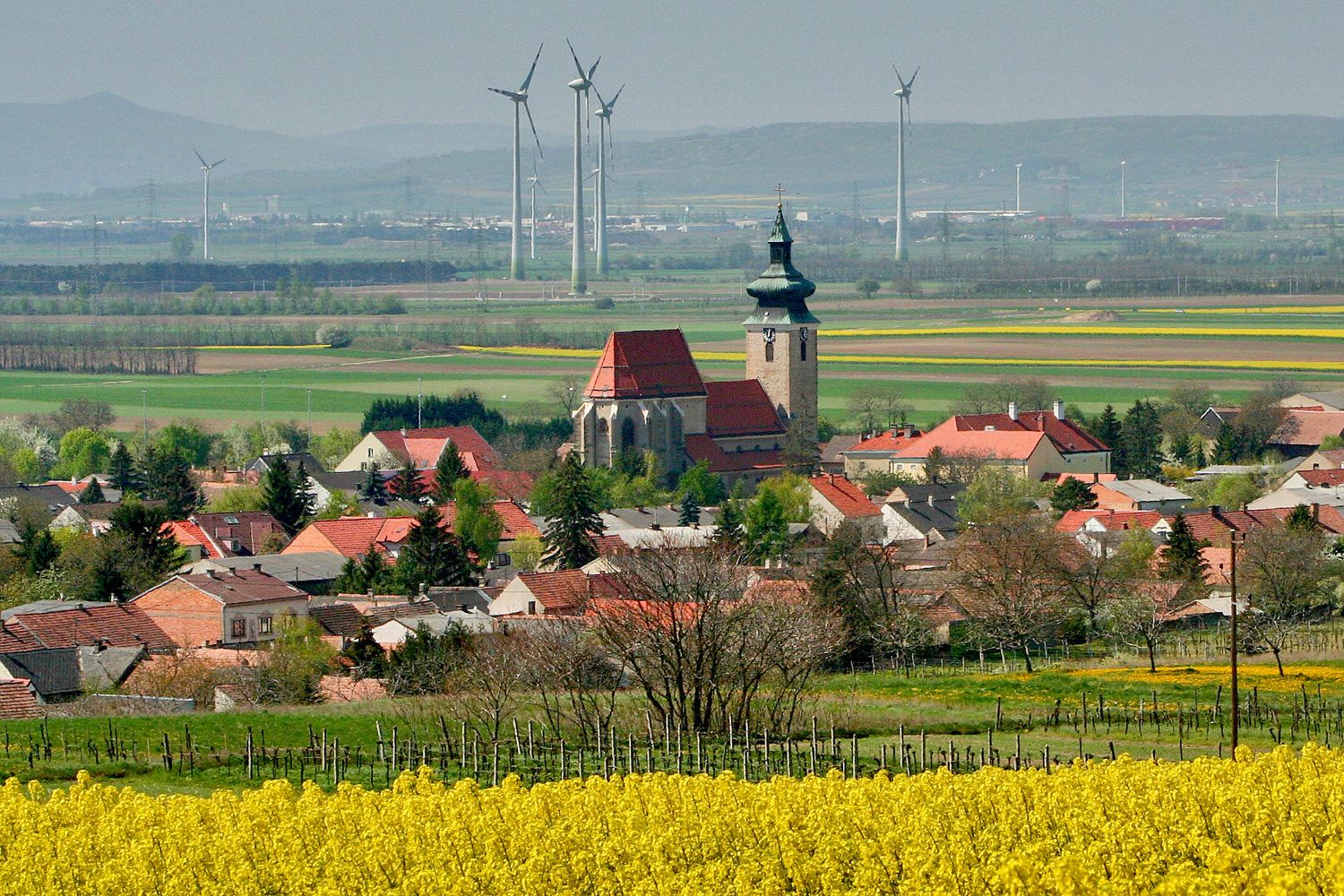View of Pillichsdorf with church, surrounded by fields and wind turbines in the background.