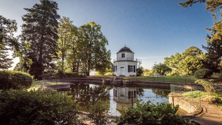 A round pavilion stands on the banks of a small pond, surrounded by trees and a well-tended garden.