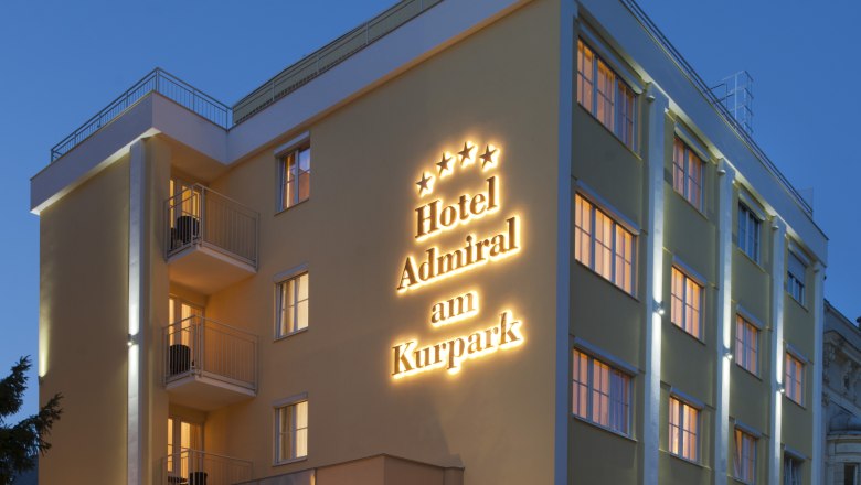 Exterior view of a hotel at dusk with illuminated lettering "Hotel Admiral am Kurpark".