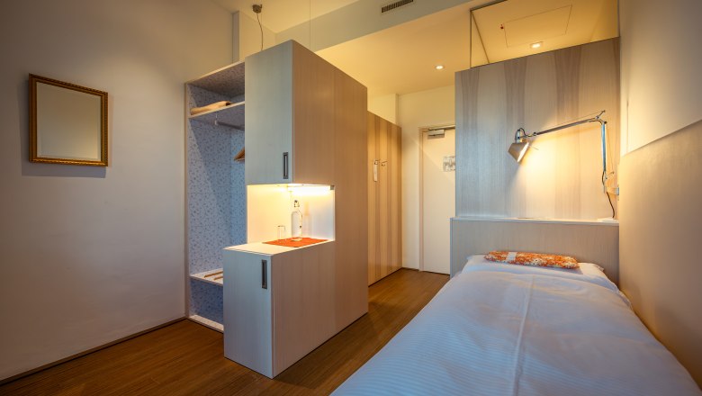Modern, minimalist hotel room with single bed, wooden floor and desk lamp.