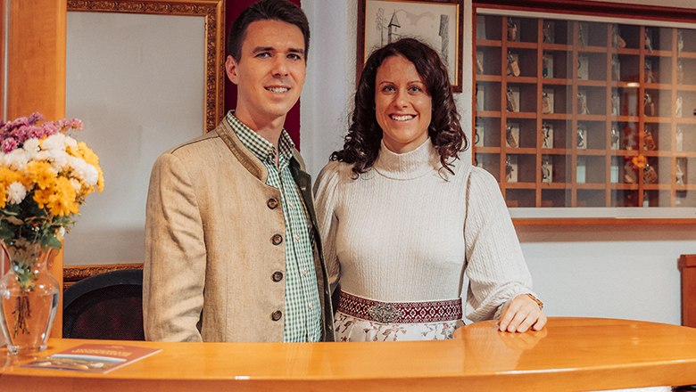 A man and a woman stand smiling behind a reception counter in traditional dress.