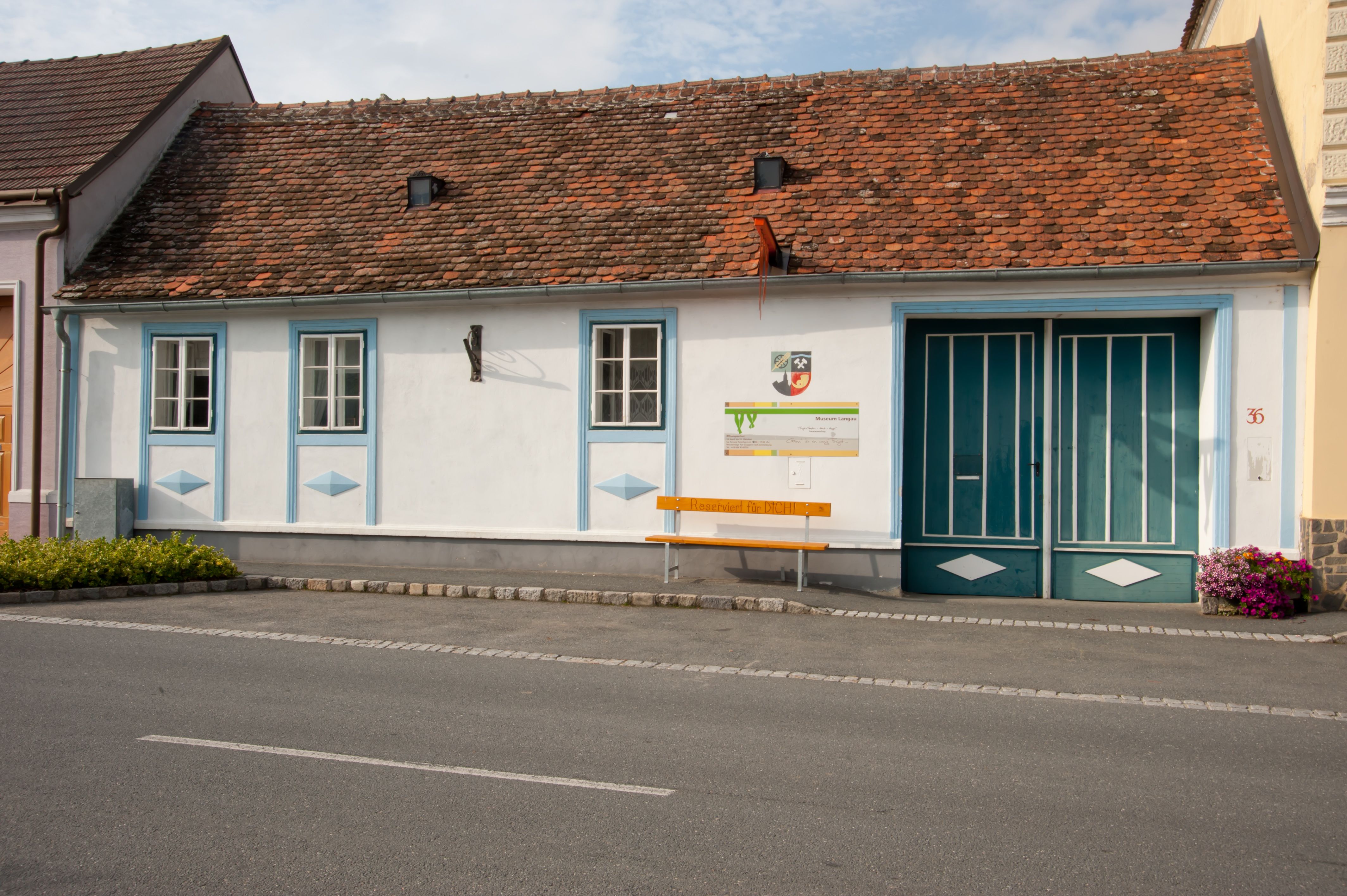 A small, traditional building with red bricks and blue window frames. In front of the building is a yellow bench with the inscription 'Reserved for YOU!