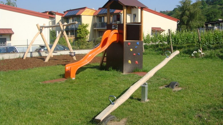 A children's playground with a slide, swing and seesaw on a meadow in front of residential buildings.