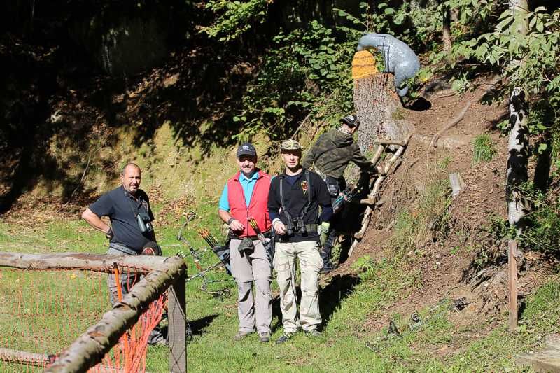 Three men with bows in the forest, one man climbing up a ladder.