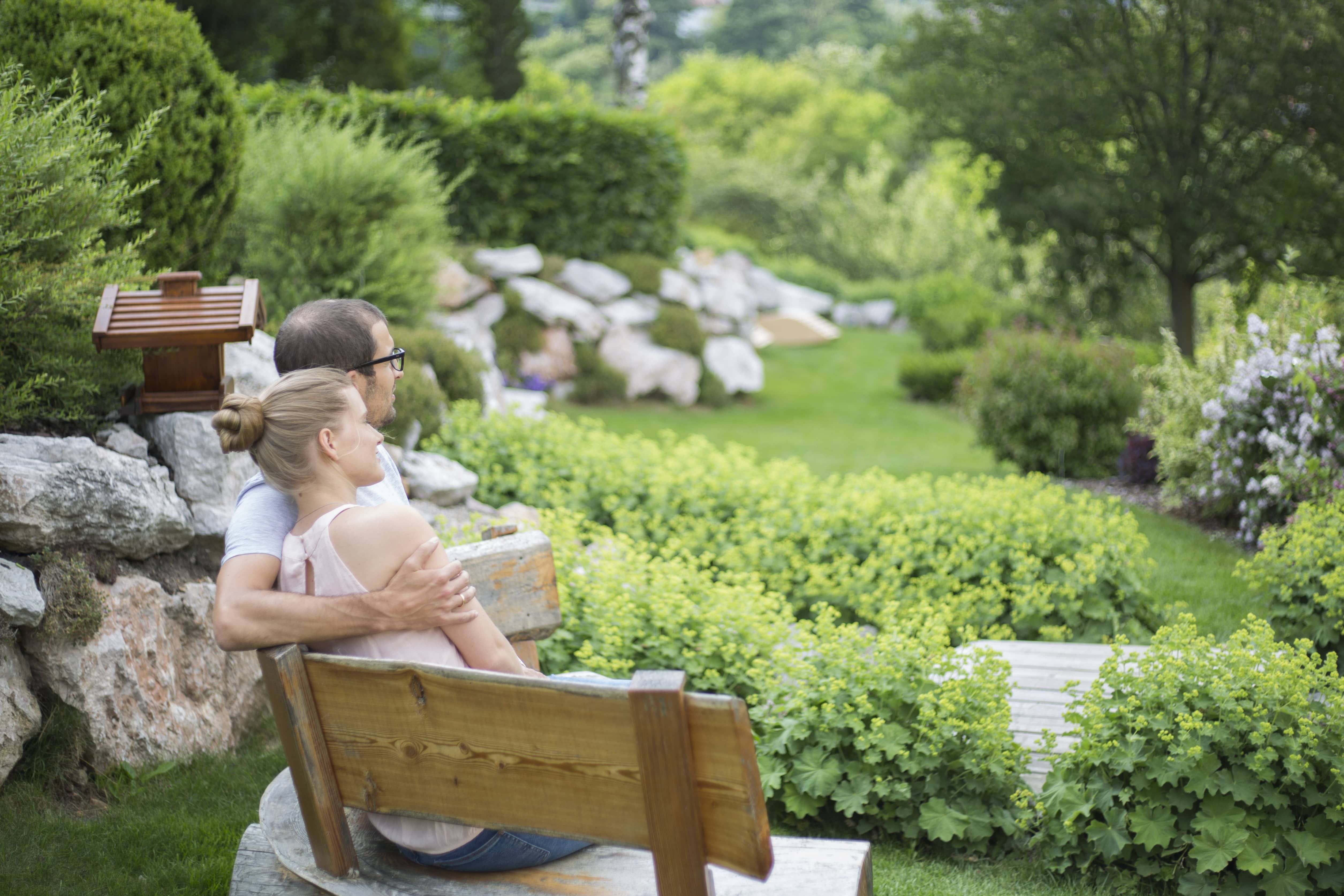 A couple is sitting on a wooden bench in a green garden with bushes and trees.