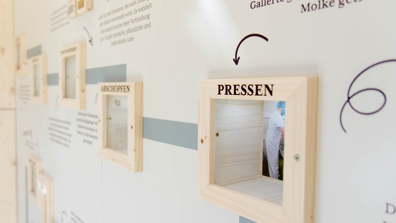 A white wall with lettering and wooden frames through which you can look into the production area of a cheese dairy.