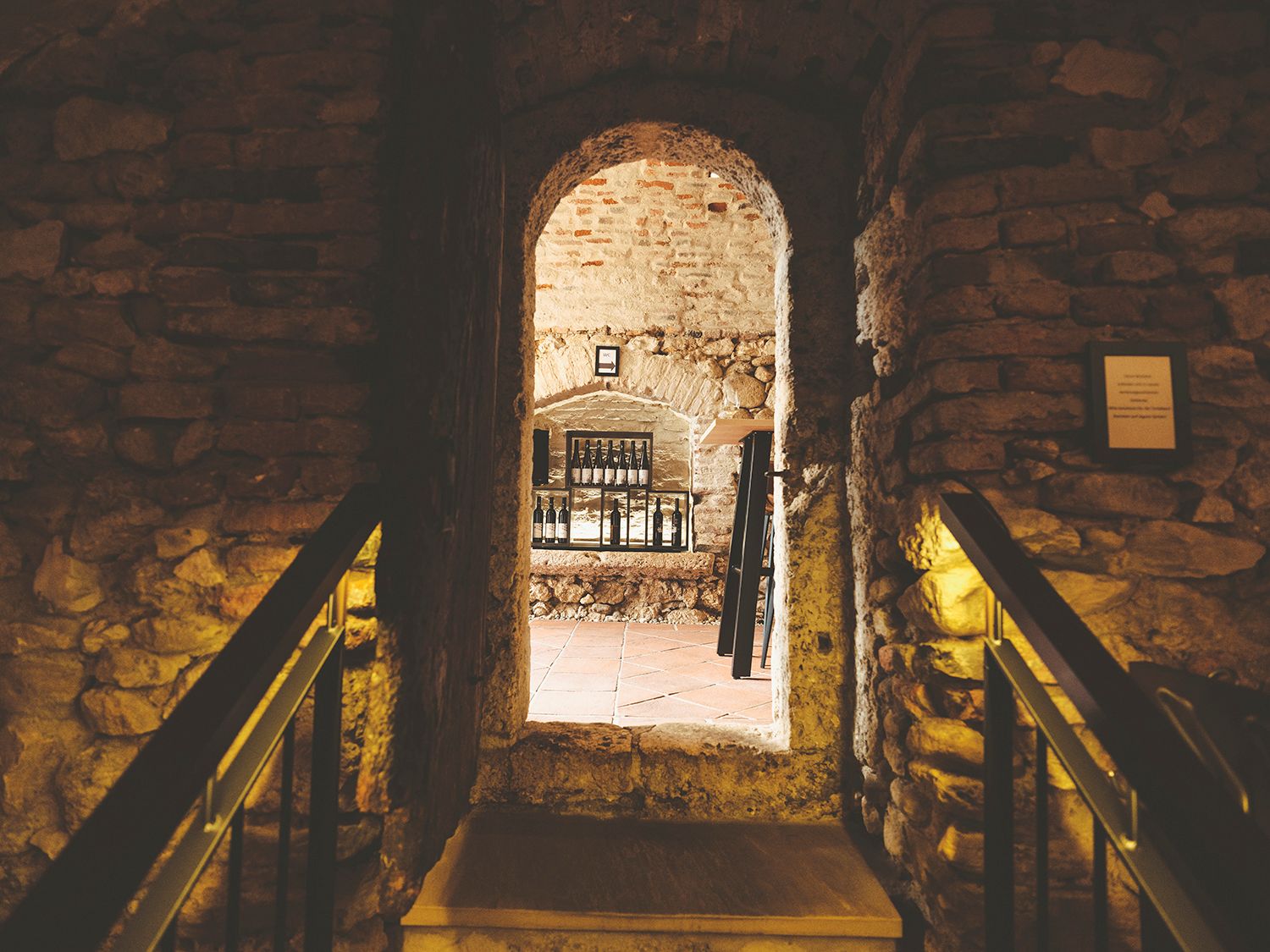Stone wall with passageway to a room with bottles on a shelf.