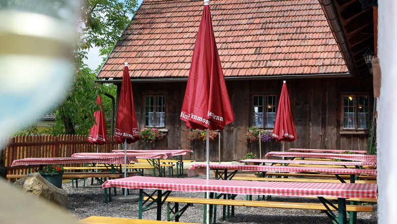 A traditional wine tavern with wooden tables, red and white checkered tablecloths and red umbrellas in front of a rustic building with a tiled roof.