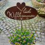 Metal sign with 'Welcome' above a pot of yellow-purple flowers on a cobbled path.