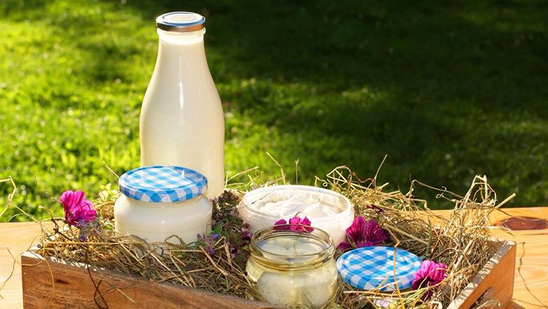 A bottle of milk and glasses of dairy products on hay in a wooden box, decorated with flowers.