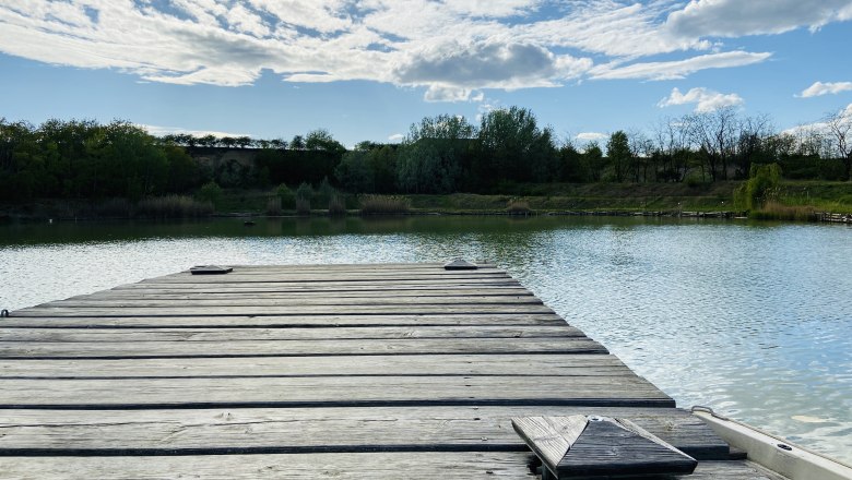 Wooden footbridge at the brick kiln pond in Zellerndorf, surrounded by trees and a blue sky with clouds.
